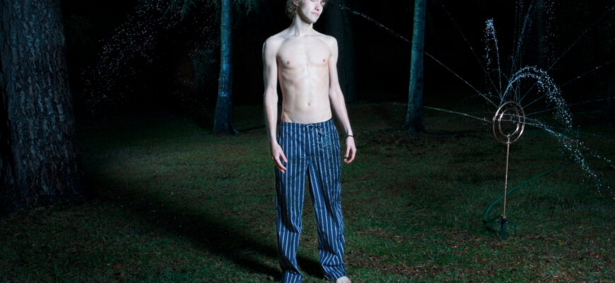 Young man standing in garden by sprinkler, eyes closed, night